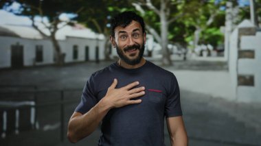 Dışarıda güneşli bir sokakta duran sakallı bir adam göğsünde bir el ile şaşkınlığını ifade ediyor.