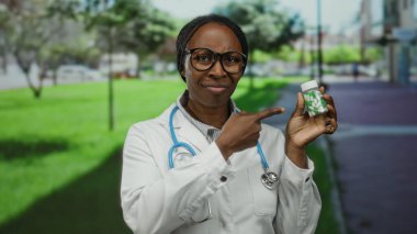 Parkta gözlüklü ve steteskoplu beyaz önlüklü bir kadın yeşil ve açık hava ile çevrili bir ilaç şişesini tutuyor ve işaret ediyor..
