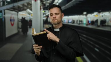 Siyah cüppeli bir adam kitap tutuyor ve arka planı bulanık tren istasyonunda kafa yoruyor..