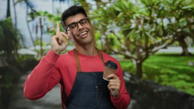 Gözlüklü, kırmızı ve önlüklü, elinde kahve fincanı tutan genç adam, gülümsüyor ve yemyeşil bir arka planda yukarı doğru bakıyor..