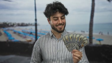 Sakallı genç adam bize dolar tutuyor, kumsalda şemsiyelerle gülümsüyor, açık deniz ortamında mutluluğu ve finansal başarıyı gösteriyor..