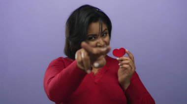 Kırmızı kalbi tutan ve mor arka plana karşı jest yapan genç bir kadın, kendine güvenen bir gülümsemeyle sevgisini ve dostluğunu ifade ediyor..