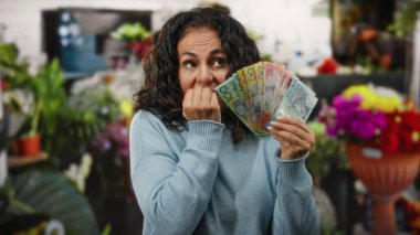 Kadın gergin bir şekilde elinde bir çiçekçi dükkanında, canlı çiçeklerle çevrili bir süveter giyerek dinamik ama endişeli bir iç mekan sahnesi yaratıyor..