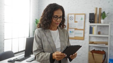 Ofiste çalışan bir kadın, elinde tabletle iyi aydınlatılmış, modern bir iş yerinde, içinde raflar ve organizasyonel materyallerin bulunduğu bir masanın yanında dururken,