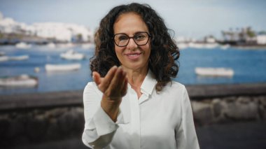 Kıvırcık saçlı bir kadın güneşli bir günde arka planda yüzen teknelerle deniz kenarındaki bir limanda el kamerasına uzanır..