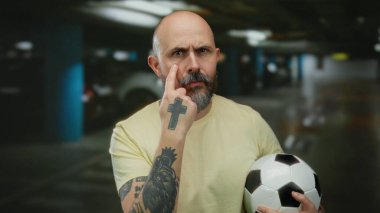Kapalı otoparkta futbol topu tutan sakallı kel bir adam ciddi bir ifadeyle işaret ediyor ve kolunda dövmesi var..