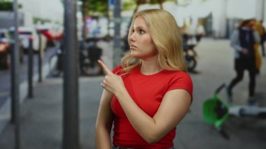 Kırmızı tişörtlü genç sarışın kadın arka planda bulanık yayalar ve bisikletlerle işlek şehir caddesinde el kol hareketi yapıyor, açık havada yön tarif ediyor..