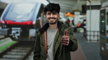 İspanyol bir adam tren istasyonunda gülümsüyor, olumlu bir jest yapıyor, modern bir demiryolu ve arka plandaki yolcularla pozitiflik ve seyahat hazırlığını iletiyor..