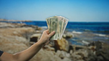 Güneşli bir sahilde Amerikan doları tutan adam, arka planda su olan deniz kenarındaki özgürlük ve finansal başarı kavramını vurguluyor..