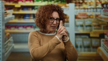 Orta yaşlı İspanyol bir kadın süpermarkette el tabancası jesti yapıyor. Etrafı raflar ve ürünlerle çevrili, gözlüklü ve kahverengi kazaklı..