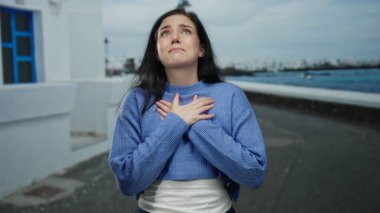 Sahilde duygularını ifade eden bir kadın mavi kazak giyiyor, yoğun duygular yakalıyor, kumral saçlar, deniz kenarı kentlerinde kendini kucaklıyor, sakin ama duygusal bir sahne.