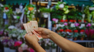 Dışarıdaki bir çiçekçide Kolombiya pezosu tutan bir adamın elleri, dış ortamda hareketli bir iş anlaşması olduğunu gösteriyor..