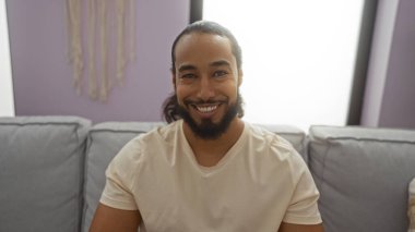 Oturma odasındaki rahat koltukta oturan gülümseyen adam pastel duvarlar ve yumuşak ışıklandırmayla sıcak, davetkar bir atmosfer yaratıyor. Rahatlama ve rahatlama anı yakalıyor..