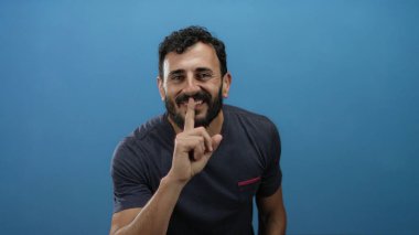 Sakallı İspanyol bir adam parmaklarını dudaklarına koyuyor. Mavi arka planda sessizlik ima ediyor..