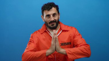 Turuncu hapishane tulumu giyen İspanyol sakallı adam, sakin ve içsel bir duayı gösteren mavi bir arkaplanda poz veriyor..