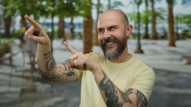 Sakallı bir adam açık havada gülümserken şehir parkı ortamını işaret ediyor, kel, modaya uygun, anlamlı jestler, gündüz, yaz, rahat bir atmosfer..