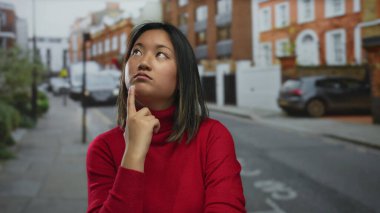 Kırmızı kazak giyen genç kadın şehir manzarasının bulanık arka planına ve park edilmiş arabalara sahip şehir sokaklarında özenle poz veriyor..