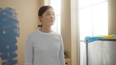 Yeni dekore edilmiş oturma odasında elinde boya malzemeleriyle duran bir kadın. Ev ortamında çekilmiş. Asyalı estetik ve modern bir ev atmosferi sergiliyor..