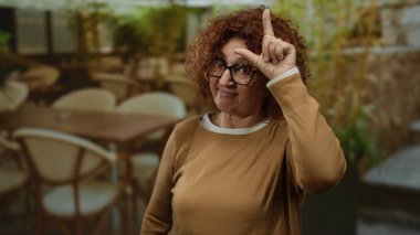 Kadın esmer orta yaşlı jestler açık hava restoranının terasında kaybedenler kahve ve sandalyelerle arka planda komik ve oyuncu ifadeleri resmediyor..