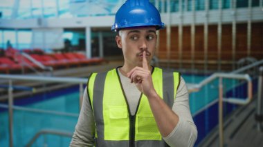 Güverte sandalyeleri ve denizle çevrili yolcu gemisinde sert şapka ve yansıtıcı yelek giyen genç adam susma hareketi yapıyor.