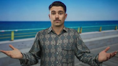 Deniz kenarında gezinen genç İspanyol bir adam berrak mavi bir gökyüzü ve okyanus arkaplanına karşı derin bir ifadeyle, sakin bir açık deniz manzarası sergiliyor..