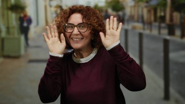 Kıvırcık saçlı İspanyol bir kadın şehir merkezinde gözlük ve açık havada mor bir süveter giyerek şakacı hareketler yapıyor..