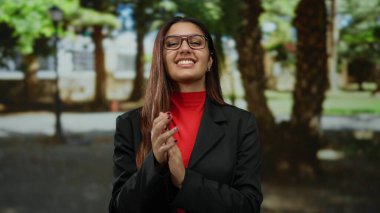 Güneşli bir park ortamında mutlu bir şekilde alkışlayan, gözlük takan, canlı kırmızı ve siyah kıyafetli bir kadın, dışarıda olumlu ve başarılı olma hissi uyandırıyor..
