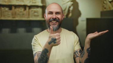 Sakallı ve dövmeli, gülümseyen kel bir adam, müze ortamında coşku ve pozitiflik gösteren başparmaklarını öne çıkarıyor..