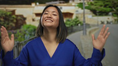 Genç bir kadın, parlak bir günde şehir caddesinde mavi bir tişört giyerek binaların ve yeşilliklerin olduğu duyguları yansıtan bir jest yapıyor..