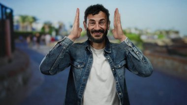 Sakallı, gülümseyen ve çerçeveli, güneşli bir sokakta açık havada kot ceket giyen İspanyol bir adam..