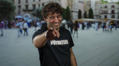 Gönüllü tişört giyen İspanyol adam işlek caddede gülümseyip el kol hareketleri yapıyor. Şehir ortamında samimi ve iç açıcı bir anı yakalıyor..