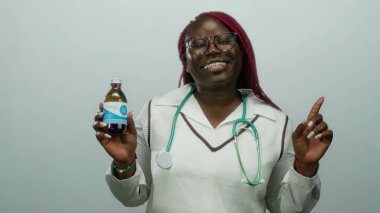 Elinde ilaç şişesi tutan gülümseyen kadın doktor steteskop profesyonel izole edilmiş beyaz duvar arkaplanı bardakları işaret ediyor.