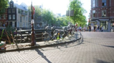 Arka planda tarihi binalar ve yapraklı ağaçlar olan güneşli bir kanal boyunca park etmiş bisikletlerin bulanık görüntüsü rüya gibi bir şehir manzarası yaratıyor..
