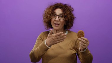 Mor arka planda kurabiye tutan orta yaşlı, kıvırcık saçlı ve gözlüklü bir kadın elinde yulaf ezmeli bisküviyle sürpriz yaparak neşeli bir sahne yaratıyor..