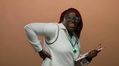 Sırt ağrısı çeken doktor pano tutuyor, kahverengi arka planda duruyor, sağlık durumundaki rahatsızlığı gösteriyor..