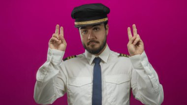 Pilot üniforması içinde sakallı genç adam pembe arka plana karşı jest yapıyor, şakacı bir şekilde alaycılık ve mizah yapıyor..