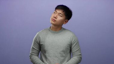 Gri kazaklı genç adam mor arka planda, sakin bir ifadeyle, gözleri kapalı ve hafif bir gülümsemeyle, huzurlu ve rahat bir ruh hali içinde duruyor..