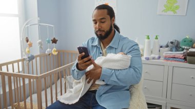 Yatak odasında oturan bir adam bebeğini kucağında tutuyor ve evdeki bebek beşiği ve şifoniyerinin yanında modern aile hayatını yansıtan mavi duvarlı akıllı telefon kullanıyor..
