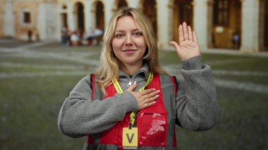 Açık hava üniversite ortamında, kırmızı yelek ve sarı saç giyen, eğitim ortamında kendini adamış ve bağlılık yemini eden bir kadın..
