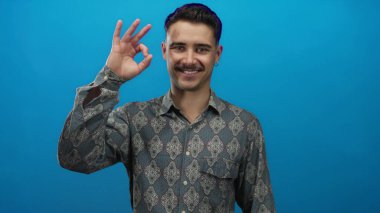 Şablonlu genç İspanyol adam canlı mavi arka planda, kendine güveni tam bir jest yaparak pozitiflik ve güvenilirliği ima ediyor..