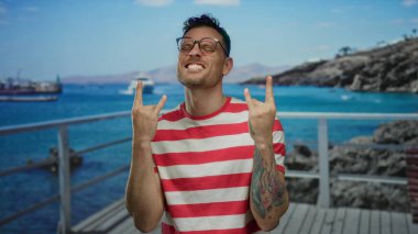Kırmızı çizgili gözlüklü genç adam kaya el işareti gösteriyor, canlı mavi okyanus arka planına sahip bir güvertede neşeyle gülümsüyor, eğlence ve özgürlüğü yakalıyor..