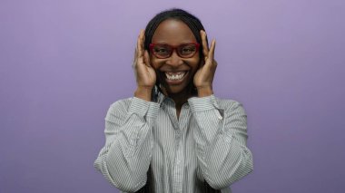 Gözlüklü, örgülü, çizgili tişörtlü, mor arka planda gülümseyen ve şakacı yüzünü saklayan bir kadın..