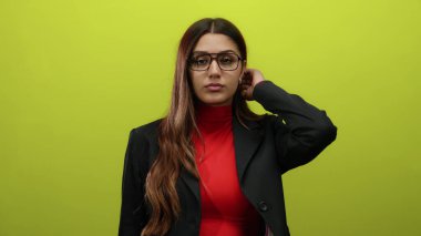 Gözlüklü ve takım elbiseli, sarı renkli bir arkaplan karşısında tek başına duran bir kadın.