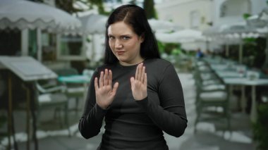 Esmer saçlı bir kadın sokak manzaralı bir restoranın terasında meraklı görünüyor. Sıradan bir şehir yemekhanesinde anlamlı bir dinleme hareketi sergiliyor..
