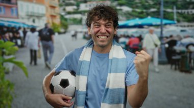 İspanyol adam futbol topu tutuyor ve atkı takıyor. Güneşli bir sokakta heyecanlı bir şekilde gülümsüyor..