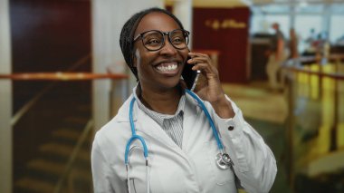 Doktor kadın otel lobisinde akıllı telefondan konuşuyor, gözlük ve laboratuvar önlüğü takıyor, steteskoplu, gülümsüyor ve mutlu bir şekilde el kol hareketi yapıyor..