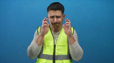Yansıtıcı yelek giyen genç İspanyol adam umut ya da batıl inanç gösteren izole edilmiş mavi arka plan duvarına karşı şans diliyor..