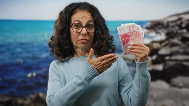 Elinde İzlanda kronu olan orta yaşlı bir kadın deniz manzarası üzerinde duruyor, sakin bir plaj zeminine karşı ekonomik temaları vurguluyor..
