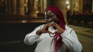 Saçları örgülü ve gözlüklü bir kadın, kilisede dikiliyor, beyaz elbiseler giyiyor, neşe ve sıcaklık ifade ediyor..