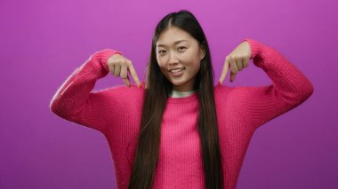 Pembe kazak giyen kadın canlı pembe duvara karşı duruyor. İki eliyle aşağıya bakıyor, oyuncu havasını ve stüdyo ortamındaki kendine güvenini yakalıyor..